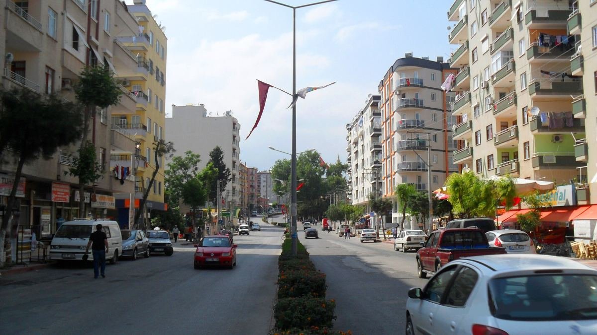 Toroslar'da iki yanı apartmanlarla çevrili, refüjlü ve ağaçlı geniş bir cadde; yolda araçlar ve yayalar var